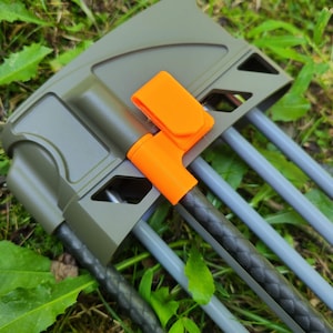 May include: An olive green archery quiver featuring a bright orange plastic clip attachment. The quiver holds several arrows with grey shafts and one arrow with a textured black carbon fiber shaft, all secured within the molded frame structure.