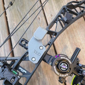 May include: A black compound bow with a gray bracket that reads "TKD". The bow features a sight with the text "Spot Hogg" and a level. The bow is set against a wooden background.