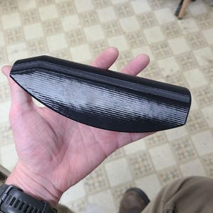 May include: A black, elongated object with a textured surface is held in a person's hand. The object appears to be made of a solid material and has a slightly curved shape. The background shows a tiled floor.