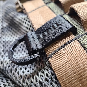 May include: Close-up of a black, 3D-printed tactical gear component with the text "IKD" visible. The component features a D-ring and is attached to tan and olive drab webbing and a gray mesh material. The image highlights the texture and details of the materials.