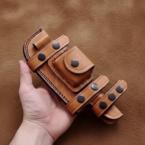 May include: A handcrafted brown leather knife sheath with multiple compartments and straps. The sheath features black snap closures and detailed stitching. The leather has a rich, warm tone, suggesting durability and quality craftsmanship.