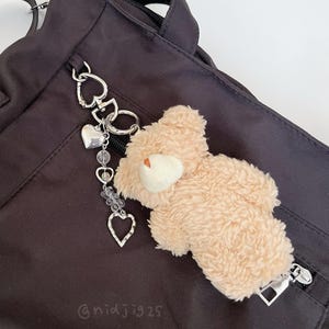 Könnte beinhalten: Ein schwarzer Rucksack mit einem silbernen Herz-Anhänger und einem kleinen Teddybär-Schlüsselanhänger. Der Anhänger hat eine Perlenkette mit klaren Perlen und silbernen Herzen.