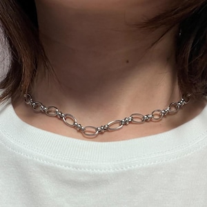 May include: A silver chain necklace with an alternating oval and circular link design. The necklace is worn on a person wearing a white t-shirt with Japanese characters and the word "AKIRA" printed on it.