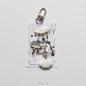 May include: A white cloud-shaped keychain with a silver charm attached. The charm features a smaller white cloud, a star, and a heart. The keychain is attached to a silver ring and a silver hook. The keychain is on a white background with a small card that says "www.etsy.com" and "NIDJIG".