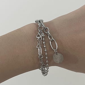 Puede incluir: Una pulsera plateada con tres hilos de diferentes diseños. Uno tiene pequeñas cuentas, otro un diseño de eslabones de cadena y el tercero una cadena con un diseño de eslabones ovalados. Un pequeño dije con un diseño de oso cuelga de la pulsera.