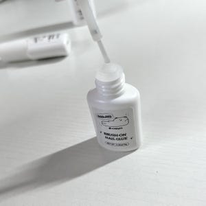 Könnte beinhalten: Eine weiße Flasche mit Pinsel-Nagelkleber mit einer Cartoon-Bärenillustration auf dem Etikett. Die Flasche trägt die Aufschrift "NIDJIG" und "BRUSH-ON NAIL GLUE".