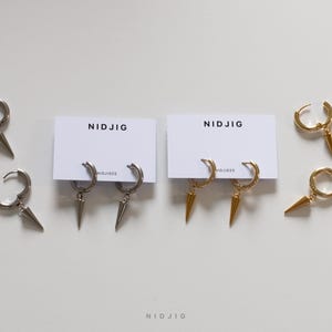 Pode incluir: Dois pares de brincos com um design pontudo. Um par é prateado e o outro é dourado. Os brincos são exibidos em cartões brancos com a palavra "NIDJIG" impressa neles.
