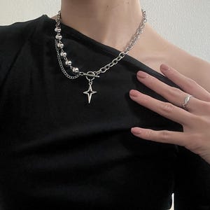 以下が含まれることがあります： 星形のペンダントとシルバービーズのチェーンが付いたシルバーチェーンネックレス。ネックレスは黒いシャツを着た人が着用しています。