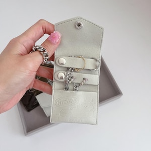 May include: A white felt jewellery case with a snap closure. The case has three compartments for storing rings, bracelets, and necklaces. The case has the word "NIDJIC" printed on the front.