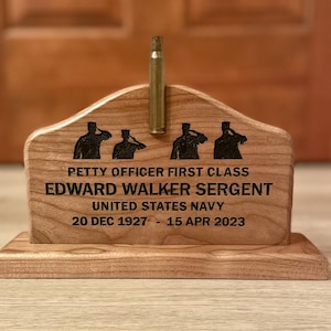 May include: A wooden memorial plaque with the text "PETTY OFFICER FIRST CLASS EDWARD WALKER SERGENT UNITED STATES NAVY 20 DEC 1927 - 15 APR 2023". The plaque features saluting figures and a brass bullet casing.
