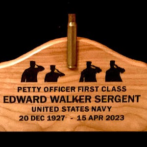 May include: A wooden plaque with the text "PETTY OFFICER FIRST CLASS EDWARD WALKER SERGENT UNITED STATES NAVY 20 DEC 1927 - 15 APR 2023". The plaque features silhouettes of saluting figures and a bullet casing.