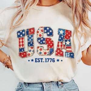 Könnte beinhalten: Cremefarbenes T-Shirt mit dem Wort "USA" in Patchwork-Buchstaben, mit roten, weißen und blauen Mustern. Unter den Buchstaben steht der Text "EST. 1776" mit Sternen auf beiden Seiten. Das Shirt hat ein lässiges, patriotisches Thema.