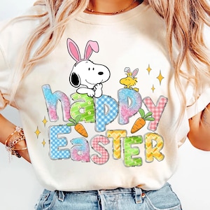 Könnte beinhalten: Ein cremefarbenes T-Shirt mit den Worten "Happy Easter" in bunten, gemusterten Buchstaben. Der Buchstabe "h" zeigt Snoopy mit Hasenohren. Ein gelber Vogel mit Hasenohren ist ebenfalls vorhanden, zusammen mit Karottenillustrationen und Sternakzenten.