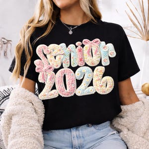 Könnte beinhalten: Schwarzes T-Shirt mit dem Text "Senior 2026" in einem Blumen- und Schleifendesign. Der Text ist in Pastellfarben gehalten, darunter Rosa, Blau und Gelb. Das Shirt ist ein lässiger Stil.