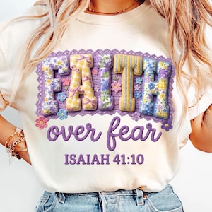Könnte beinhalten: Cremefarbenes T-Shirt mit dem gestickten Schriftzug "FAITH over fear". Das Wort "FAITH" ist in einem 3D-Blumen- und Karomuster gehalten. Der Text "ISAIAH 41:10" steht darunter in lila Schrift.