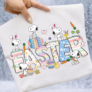 以下が含まれることがあります： カラフルな模様の文字で「EASTER」と書かれた白いスウェットシャツ。ウサギの耳とイースターエッグをつけたスヌーピーのキャラクターがデザインを飾っています。ニンジンと花も含まれています。