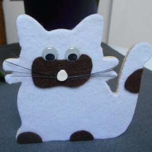 Peut inclure: Un chat en feutre blanc avec des oreilles, un nez et des pattes en feutre brun. Le chat a des yeux globuleux et un nez en feutre blanc.