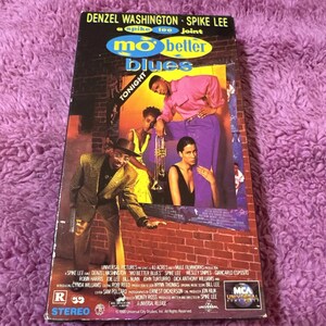 May include: A VHS tape cover for the movie "Mo' Better Blues" starring Denzel Washington and directed by Spike Lee. The cover features a red brick wall background with a group of people in the foreground. The title of the movie is in large, bold letters and the tagline "A Spike Lee Joint" is below it.