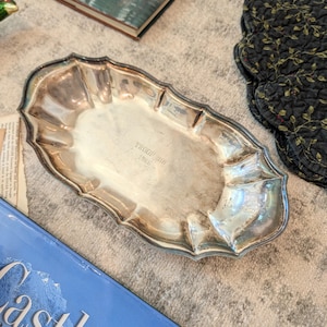 May include: A silver-colored, oval-shaped tray with a scalloped edge. The tray has the engraved text "TROOP 318 1966" in the center. The tray is on a textured surface with other items in the background.