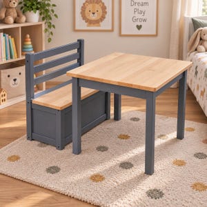 Op de afbeelding: Een kindermeubelset bestaande uit een vierkante houten tafel met een natuurlijk lichthouten blad en donkergrijze poten, gecombineerd met een bijpassende bank met een lattenrugleuning en een opbergvak onder de houten zitting.