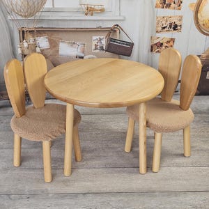May include: A set of two wooden bunny-shaped chairs with beige cushions and a round wooden table. The chairs and table are made of light wood and have a natural finish.