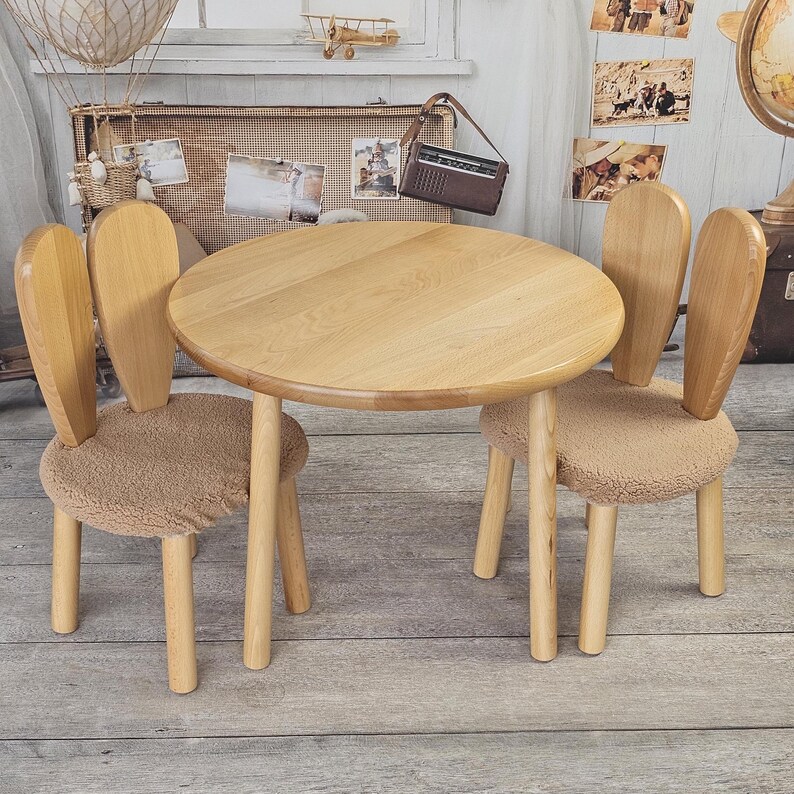 May include: A set of two wooden bunny-shaped chairs with beige cushions and a round wooden table. The chairs and table are made of light wood and have a natural finish.