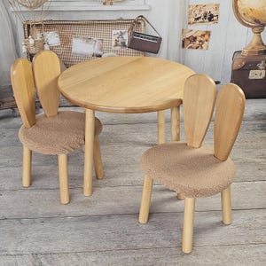 May include: A wooden children's table and two chairs with bunny ear backs. The chairs have brown fluffy cushions. The table and chairs are made of light wood.