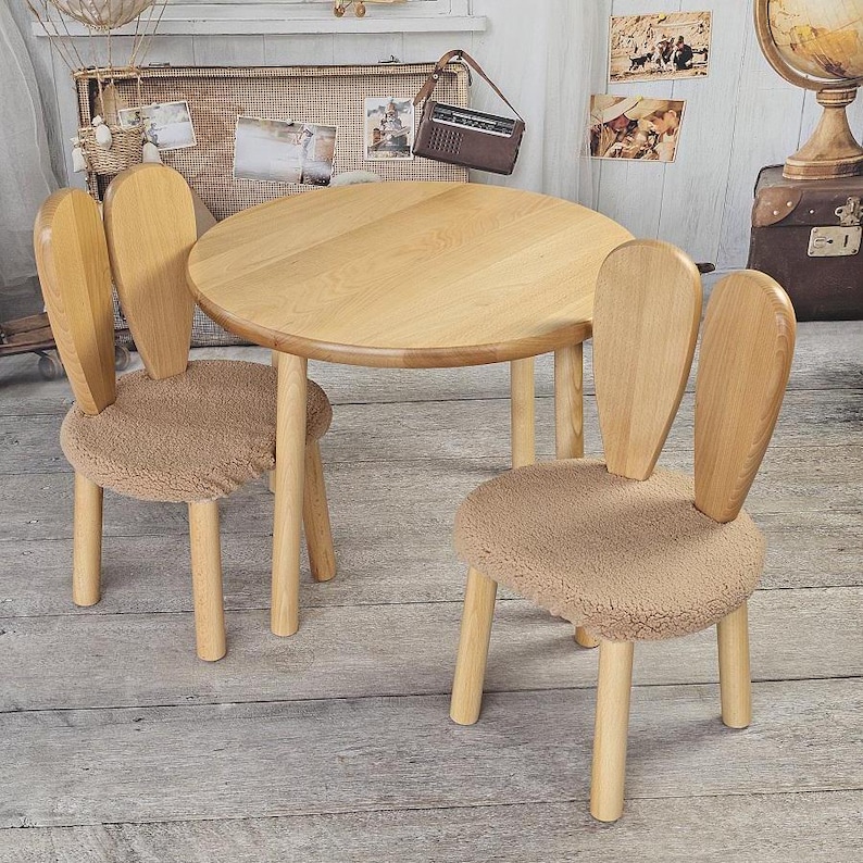 May include: A wooden children's table and two chairs with bunny ear backs. The chairs have brown fluffy cushions. The table and chairs are made of light wood.