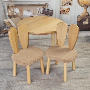 May include: Two wooden chairs with bunny ear backs and brown faux fur seats, and a round wooden table. The chairs and table are in a room with a wooden floor.