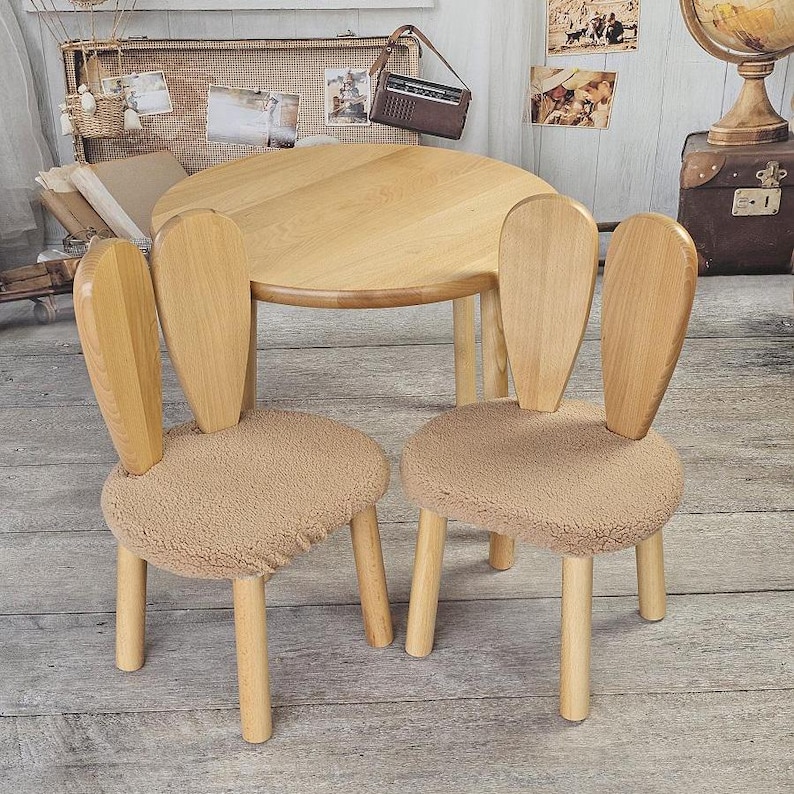May include: Two wooden chairs with bunny ear backs and brown faux fur seats, and a round wooden table. The chairs and table are in a room with a wooden floor.