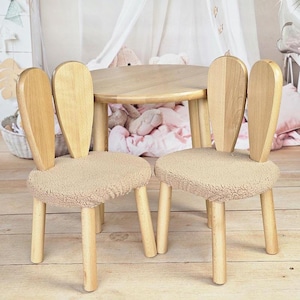 May include: Two wooden chairs with bunny ear backs and beige fluffy cushions, surrounding a round wooden table. The chairs and table are in a light wood tone.