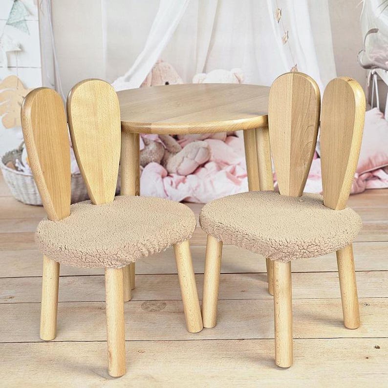 May include: Two wooden chairs with bunny ear backs and beige fluffy cushions, surrounding a round wooden table. The chairs and table are in a light wood tone.