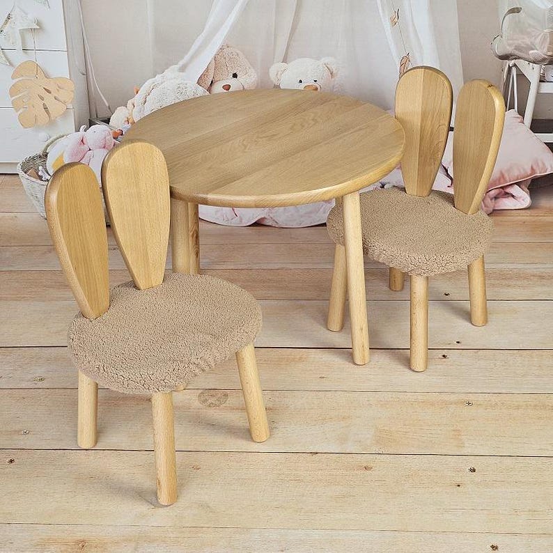 May include: A wooden table and two chairs shaped like bunny ears. The table and chairs are a light brown colour and have a soft, fuzzy beige cushion.