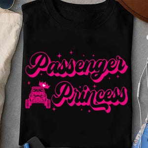 May include: Black t-shirt with the text "Passenger Princess" in pink, stylized script. The design includes a pink off-road vehicle and a small crown. Small pink stars are scattered around the text.