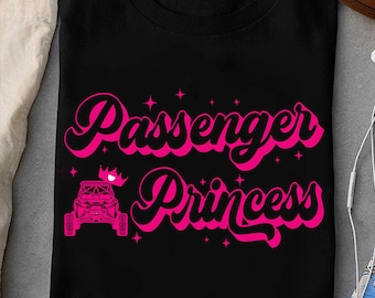 May include: Black t-shirt featuring the text "Passenger Princess" in pink, cursive lettering. The design includes a pink off-road vehicle and a small crown. Small pink stars are scattered around the text.