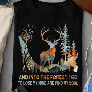 May include: Black t-shirt featuring a watercolor design of a deer with large antlers, trees, and a compass. The design includes orange, blue, and brown hues. The text at the bottom reads, "AND INTO THE FOREST I GO TO LOSE MY MIND AND FIND MY SOUL."