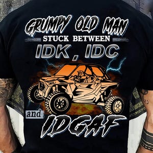 Könnte beinhalten: Schwarzes T-Shirt mit einer Grafik eines orangefarbenen Geländewagens und dem Text "GRUMPY OLD MAN STUCK BETWEEN IDK, IDC and IDGAF". Das Design umfasst Blitze und einen feurigen Hintergrund.