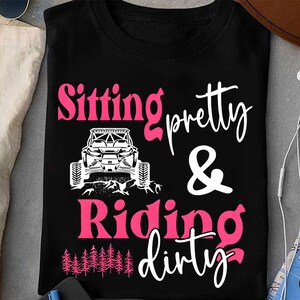 May include: Black t-shirt with the text "Sitting pretty & Riding dirty" in pink and white. A white graphic of an off-road vehicle is centered above the words "Riding dirty". The bottom of the shirt features a row of pink pine trees.