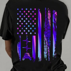 May include: Black t-shirt featuring a vibrant, stylized American flag design. The flag incorporates a heartbeat graphic, a vehicle, and stars in shades of purple, blue, and pink. The design has a distressed, textured appearance.