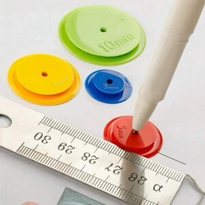 May include: A set of four circular plastic discs in green, yellow, blue, and red, each featuring a central hole and embossed size labels of 10mm, 7mm, 3mm, and 5mm. A white stylus points to the red disc, alongside a metal ruler.