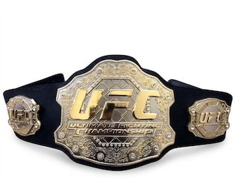 Op de afbeelding: Een zwarte kampioenschapsriem met een grote centrale goudkleurige metalen plaat met de tekst UFC ULTIMATE FIGHTING CHAMPIONSHIP, omringd door ingewikkeld filigraan en kleine heldere steentjes, geflankeerd door twee kleinere bijpassende zijplaten.