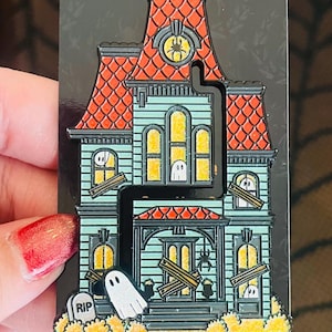 May include: A teal blue haunted house enamel pin with a red roof, boarded up windows, and a ghost in front. The pin has a black outline and glitter accents.