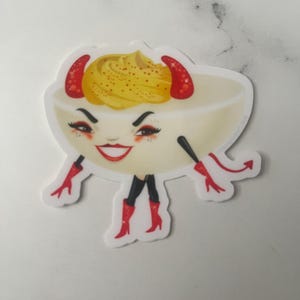May include: A die-cut sticker featuring a deviled egg character with a yellow yolk center, red horns, red gloves, black leggings, and red high-heeled boots. The character has a mischievous facial expression with dark eyebrows and red lipstick.