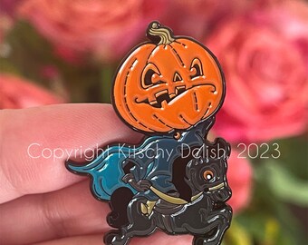 May include: A black enamel pin featuring a jack-o'-lantern riding a black horse. The jack-o'-lantern has a stitched mouth and a mischievous expression. The pin has a blue cape and the text "Copyright Kitsch Deilish 2023".