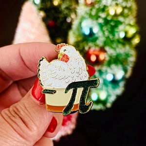 Könnte beinhalten: Eine goldfarbene Emaille-Anstecknadel mit einem weißen Huhn, das in einer Tortenkrusten sitzt, mit einem grünen Glitzer-„Pi“-Symbol. Die Anstecknadel wird in einer Hand mit rotem Nagellack gehalten.