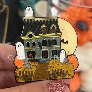 May include: A green and black enamel pin featuring a haunted house with a yellow moon and three white ghosts. The house has boarded windows and a fence with a path leading to the front door. The pin has a glittery gold finish and the text "Copyright Kitschy Design 2023".