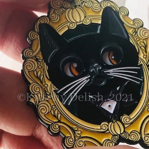 May include: Black cat with white whiskers and a bow tie in a gold ornate frame with a pumpkin design.  The text "Kissy Delish 2021" is visible at the bottom of the frame.