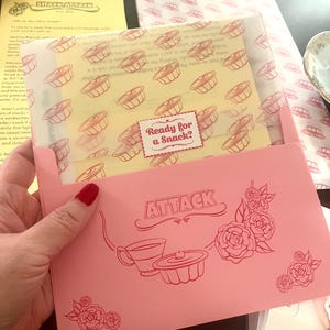 May include: A pink envelope with the word "ATTACK" and a teacup and dessert illustration. Inside, a yellow paper with a pattern of desserts and the text "Ready for a Snack?" is visible.