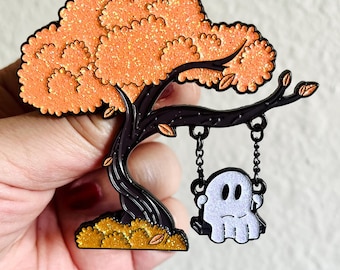 May include: A black metal enamel pin depicting a ghost on a swing hanging from a tree with orange glitter leaves. The tree trunk is black and the ground is gold glitter.