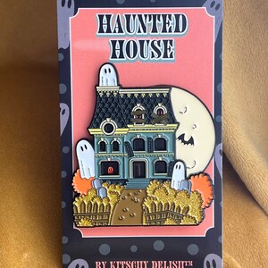 May include: A black and white enamel pin featuring a haunted house with a green door, boarded windows, and a moon with a ghost face. The house is surrounded by a fence and bushes. The pin says "Haunted House" and "By Kitschy Delish&trade;".